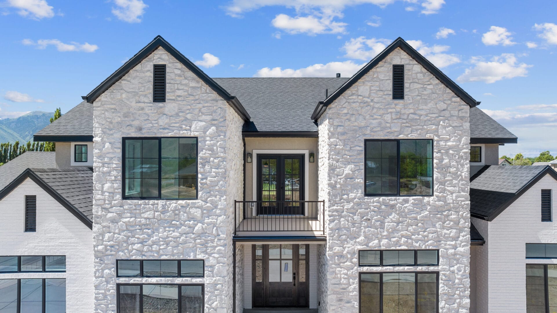 A two-story house with a stone facade, black framed windows, and a small balcony above the front entrance, set against a backdrop of blue sky and scattered clouds.