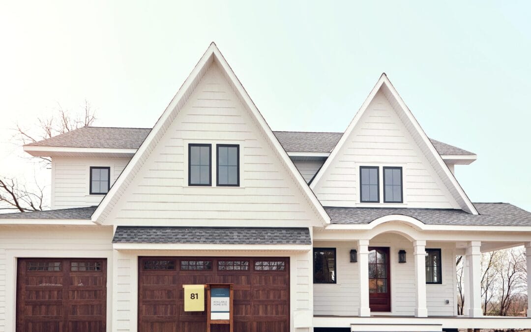 A white two-story house with three peaked roofs, black window frames, and wooden garage doors. A sign with the number 81 is displayed in front.